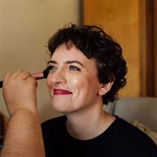 Short haired bride with bright pink lipstick smiles off camera while getting her blusher applied