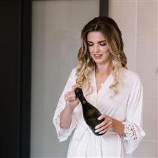 Bride with bronde waves opens prosecco bottle wearing a white robe