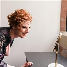 Bride in black robe with red hair and gold headband smiles at herself in a small mirror